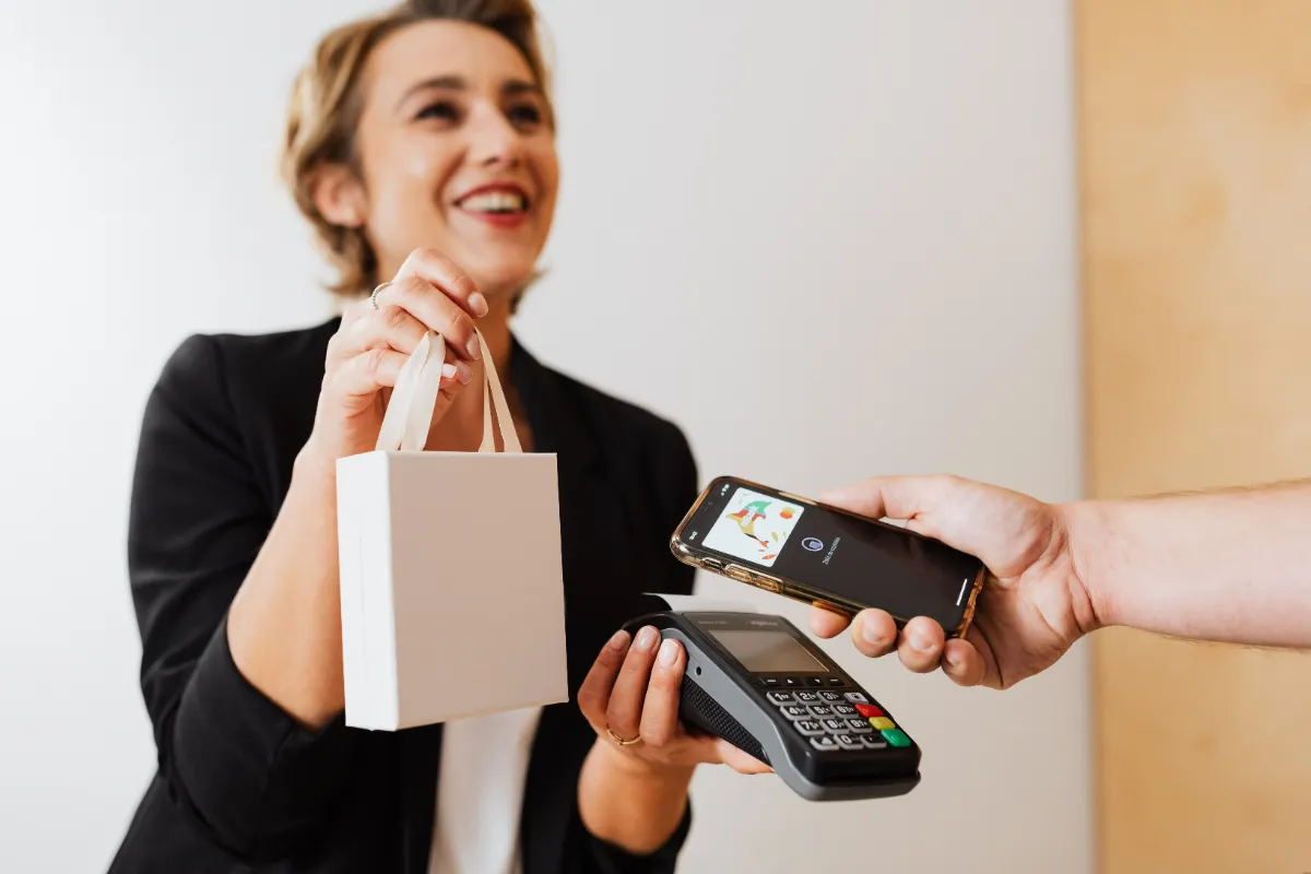 A person tapping a card against a handheld payment terminal while holding a paper bag